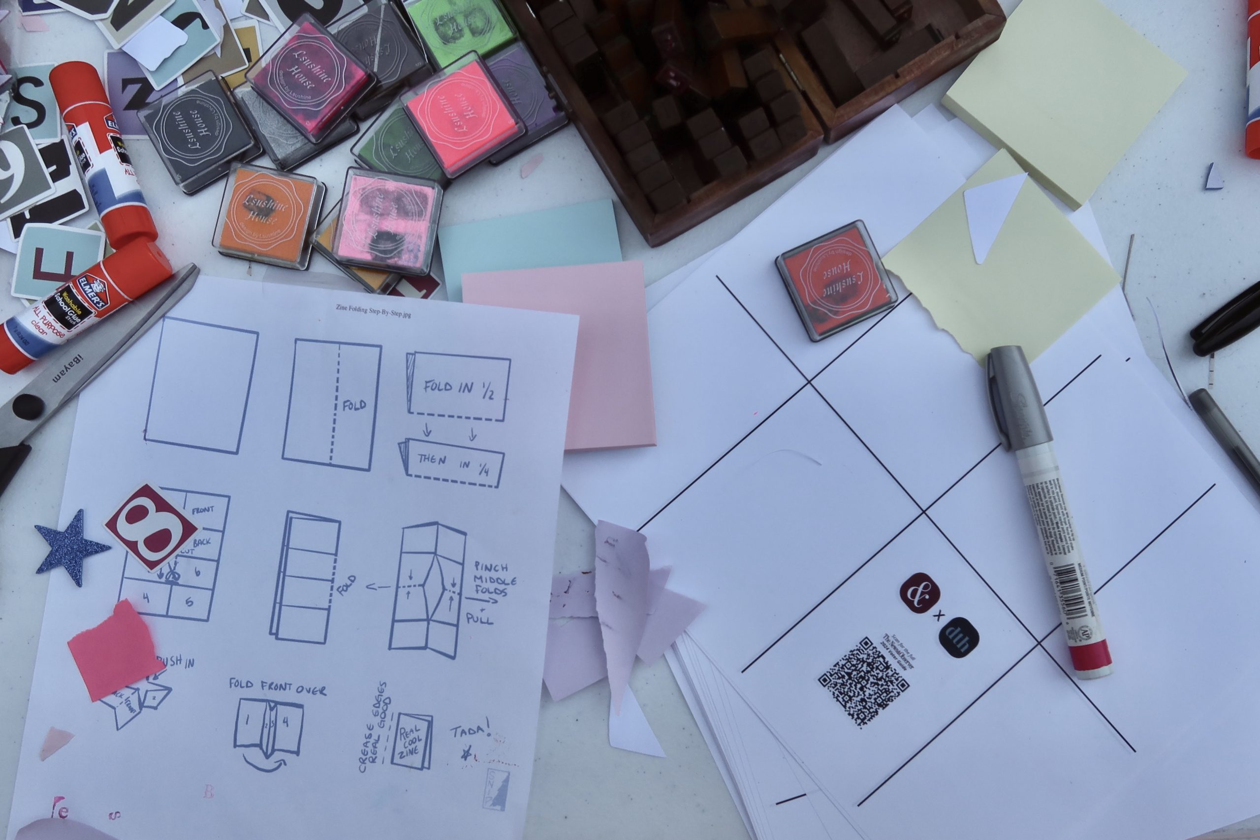 A birds-eye-view of the zine template features the N&O and DTH logos in partnership, with a QR code that is linked to the N&O’s 2024 election guide. Pictured are scattered papers with visual directions on how to fold the sheets, glue sticks, markers and other art material on a table.